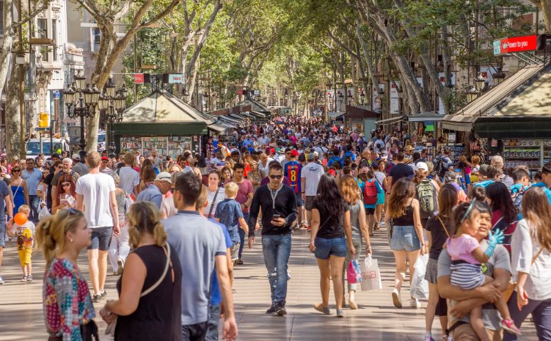 Bustling urban street with vendors, tourists, diverse crowd, trees, and shops.
