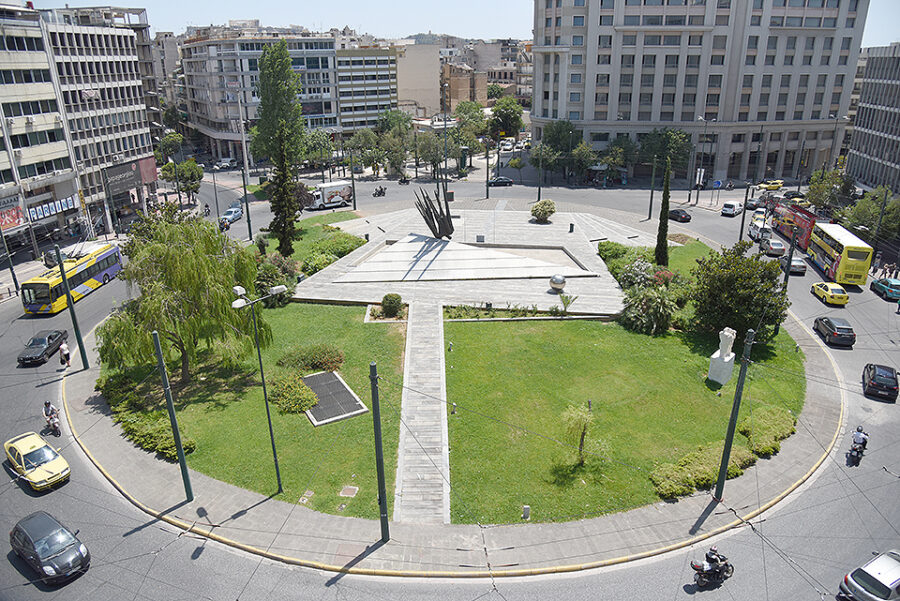 Aerial view of urban roundabout with modern sculpture, plaza, traffic and surrounding buildings.