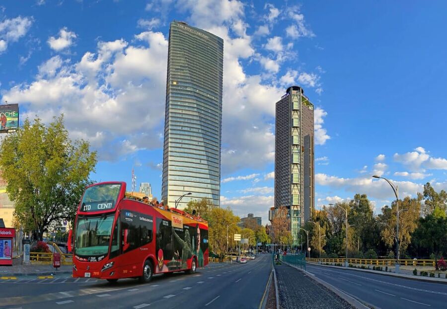 Urban scene with red bus, modern skyscrapers, greenery, and blue sky.