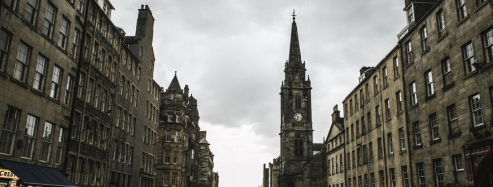 Historic European street with clock tower, bustling pedestrians, and striking architecture under stormy skies.