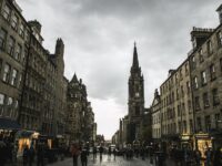 Historic European street with clock tower, bustling pedestrians, and striking architecture under stormy skies.