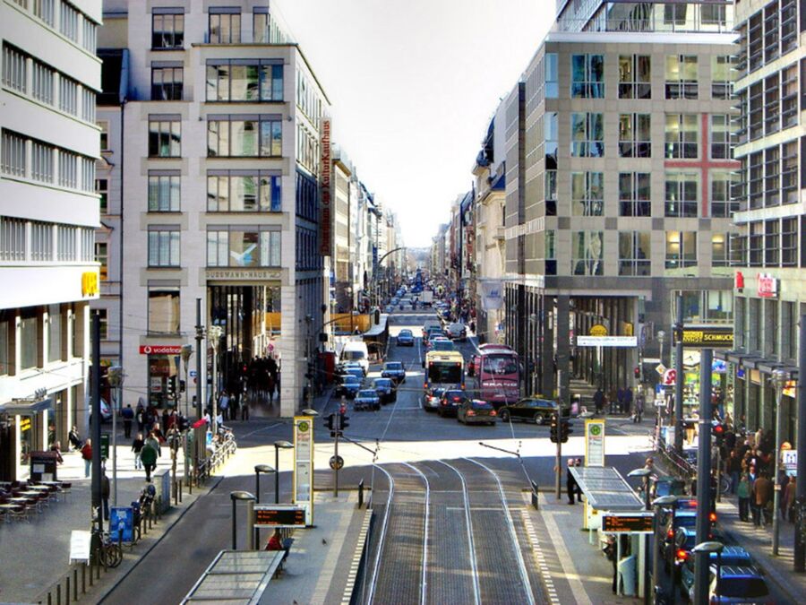Urban street with modern architecture, tram line, busy traffic, and pedestrians under sunny skies.