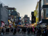 Urban shopping street at twilight with neon storefronts and bustling pedestrian crowd.