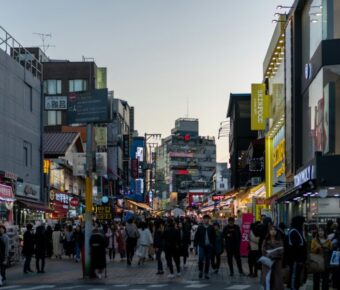 Urban shopping street at twilight with neon storefronts and bustling pedestrian crowd.