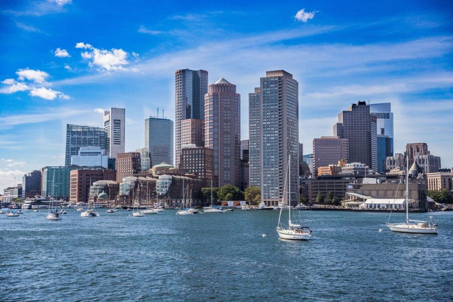 Boston cityscape featuring diverse architecture, waterfront sailboats, lush greenery, and blue skies.