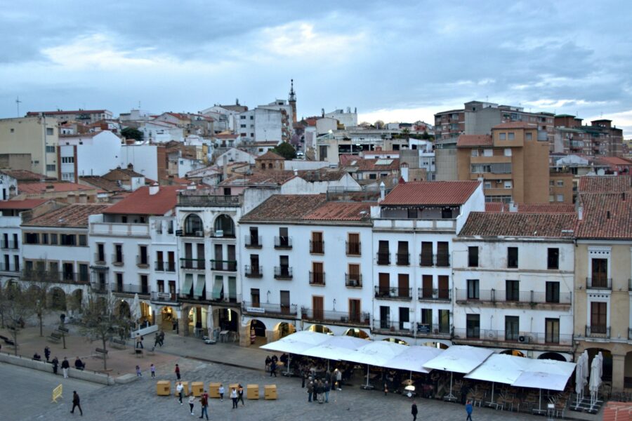 Vibrant plaza with Spanish architecture, cafes, modern buildings, and lively community gatherings.