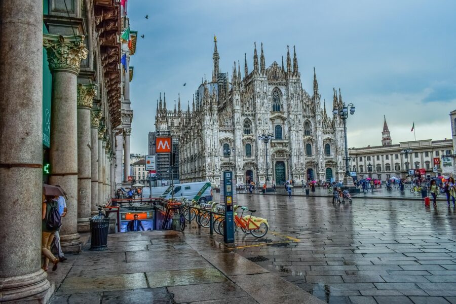 Bustling Milan Cathedral square with Gothic spires, visitors, bicycles, and metro entrance on cloudy day.