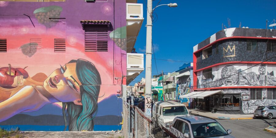 Colorful Santurce street art murals on urban buildings with cars parked along a sunny road.
