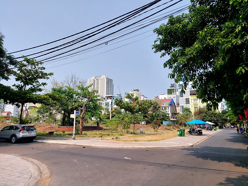 Sunny An Thuong urban street with parked car, motorbikes, trees, small park, and high‑rise buildings.