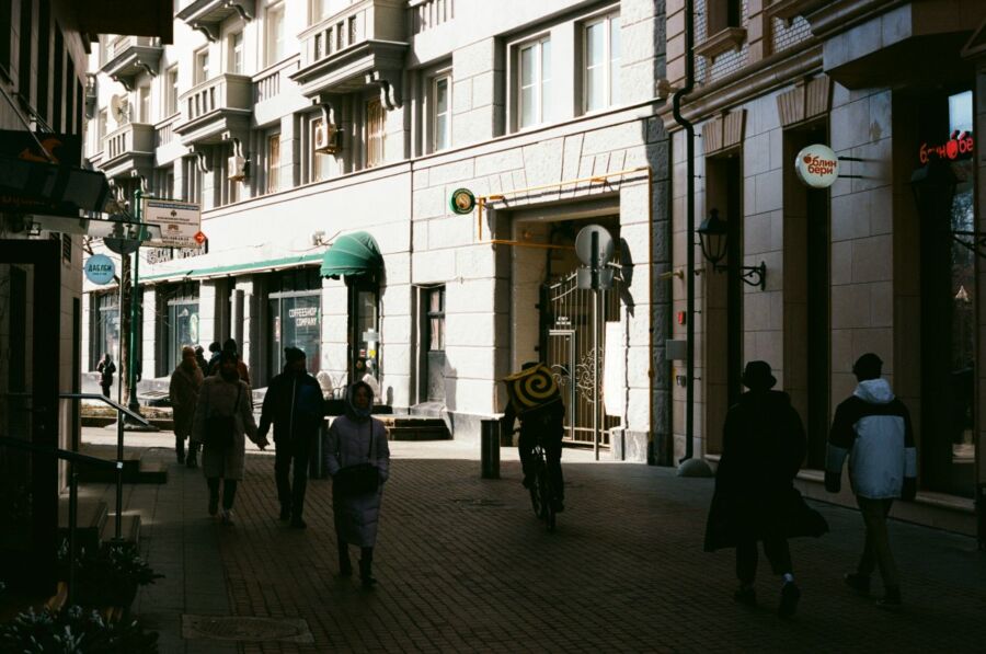 Bustling urban street scene with eclectic architecture, pedestrians, cyclist, shops, and natural elements.