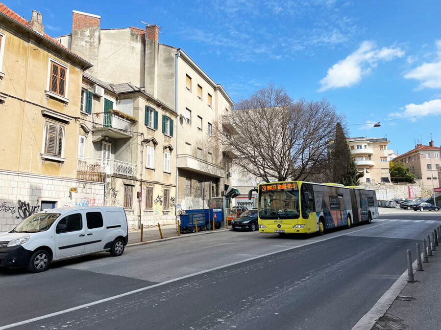 Urban street scene with old buildings, yellow bus, parked cars, and blue sky.