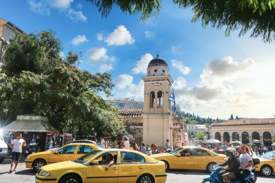 Bustling Athens square with yellow taxis, bell tower, ancient ruins, and blue skies.