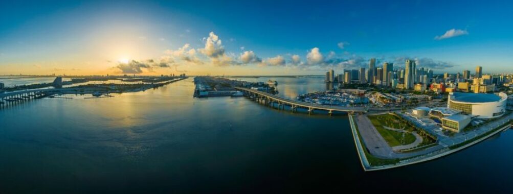 Coastal city sunrise with skyline, docks, and reflections on calm water.