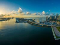Coastal city sunrise with skyline, docks, and reflections on calm water.