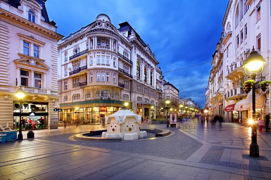 Scenic urban street at twilight with historical architecture, fountain, and storefronts on Knez Mihailova Street.