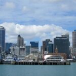 Coastal city skyline with skyscrapers, waterfront, boats; vibrant urban landscape against a bright sky.