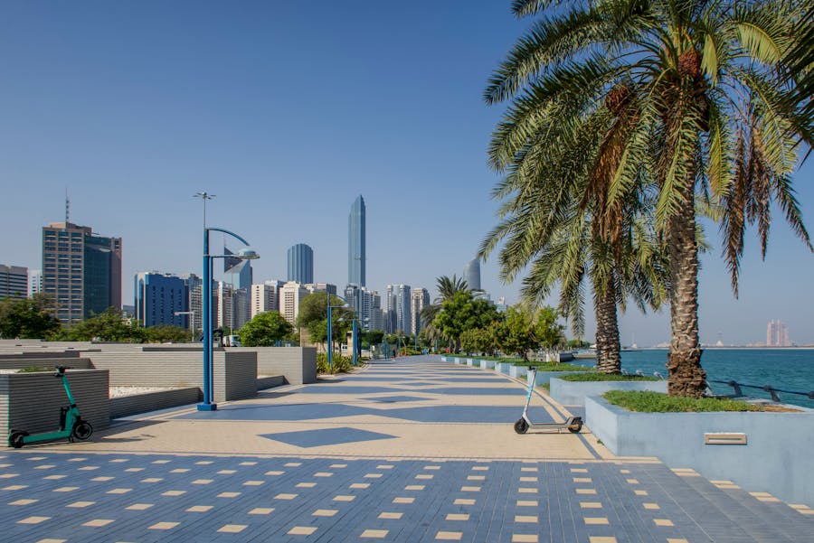 Modern waterfront promenade with skyscrapers, palm trees, and scooters under a clear blue sky.