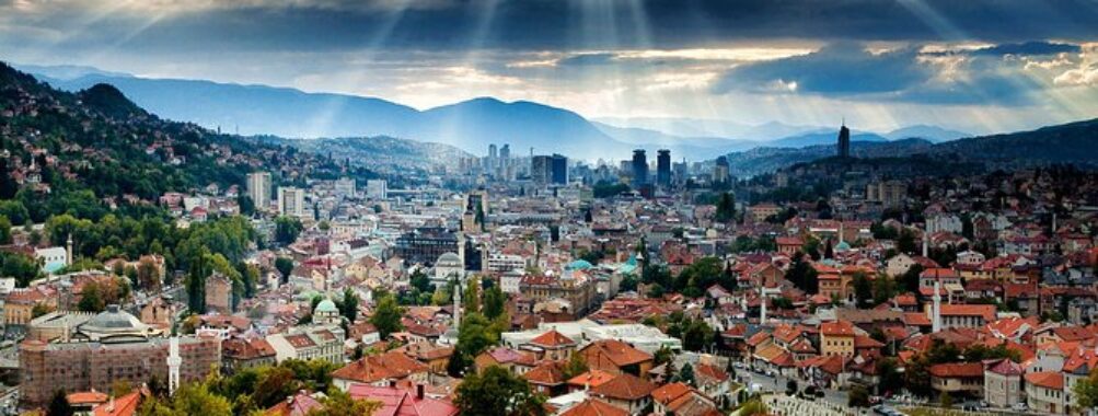 Dramatic valley city skyline with red rooftops, hillside cemetery and sun rays breaking storm clouds.