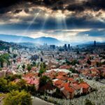 Dramatic valley city skyline with red rooftops, hillside cemetery and sun rays breaking storm clouds.