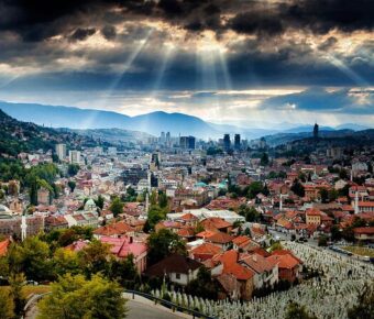 Dramatic valley city skyline with red rooftops, hillside cemetery and sun rays breaking storm clouds.