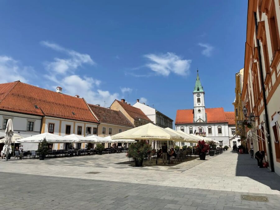 Bustling Varaždin Main Square featuring tables with umbrellas in front of a historic church.