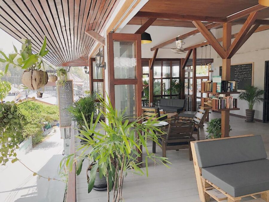 Sunlit wooden veranda café and reading lounge with plants, bookshelves, cozy seating, and tropical decor.