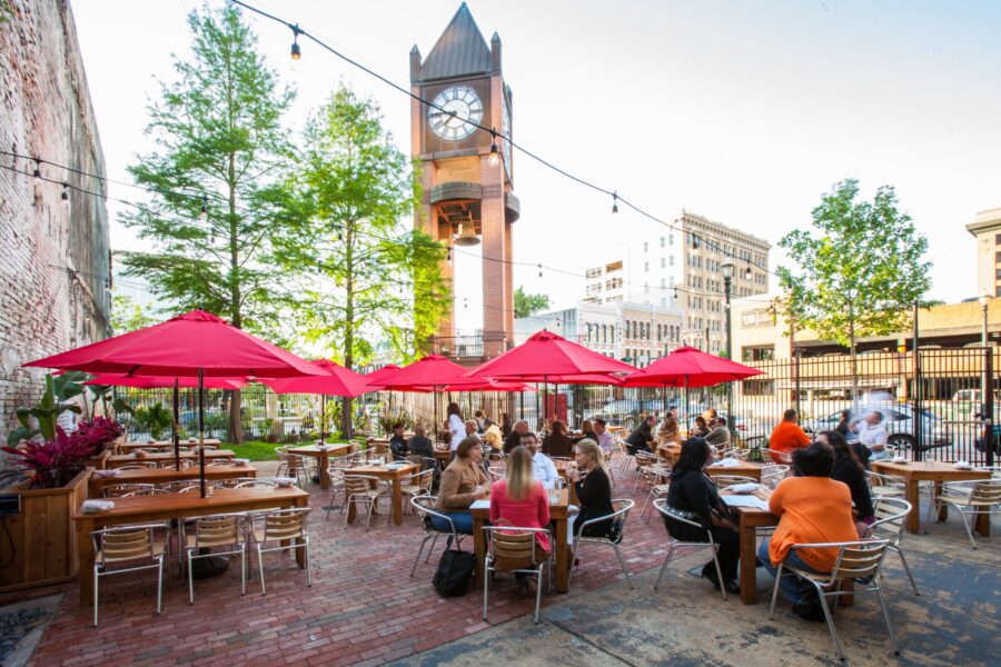 Outdoor dining scene with clock tower, red umbrellas, lush greenery, and city backdrop.