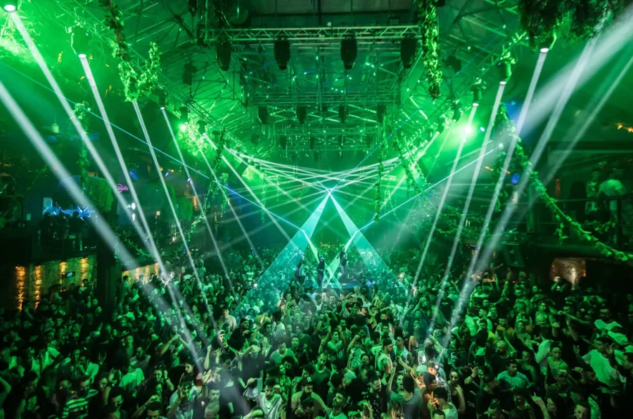 Green-lit nightclub scene with crowd dancing, laser beams, and vibrant energy.