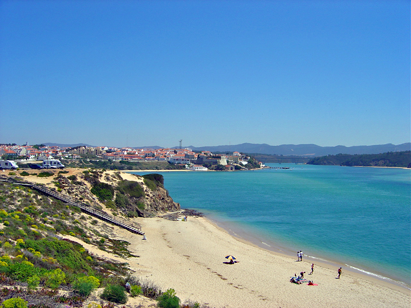 People relax on a beach in Vila Nova de Milfontes, Portugal, with a serene body of water stretching out in the background.