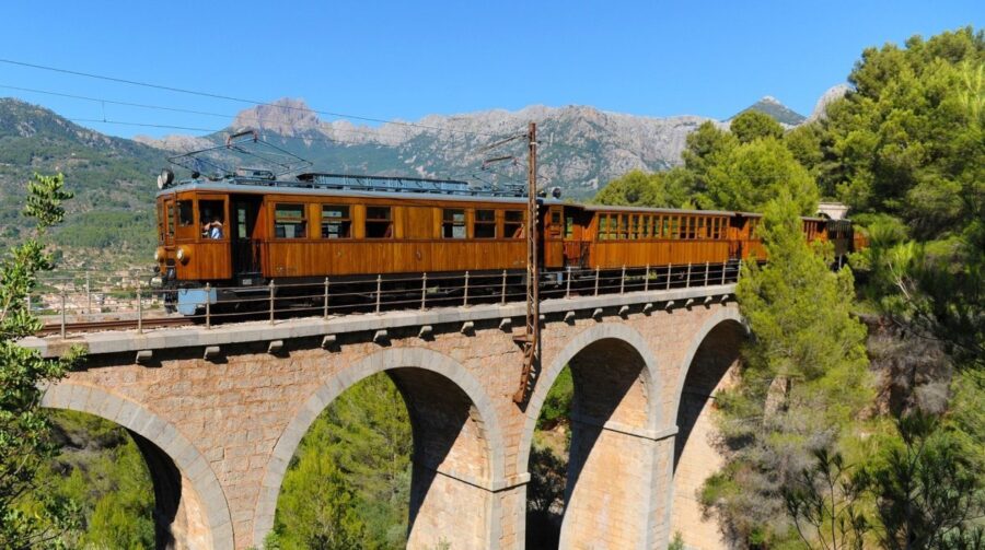 Vintage electric train crossing stone bridge with mountains, trees, and blue sky backdrop.