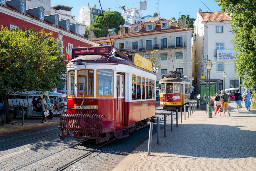 Vintage maroon tram on cobblestone street in historic Mediterranean city, tourists and market stalls.