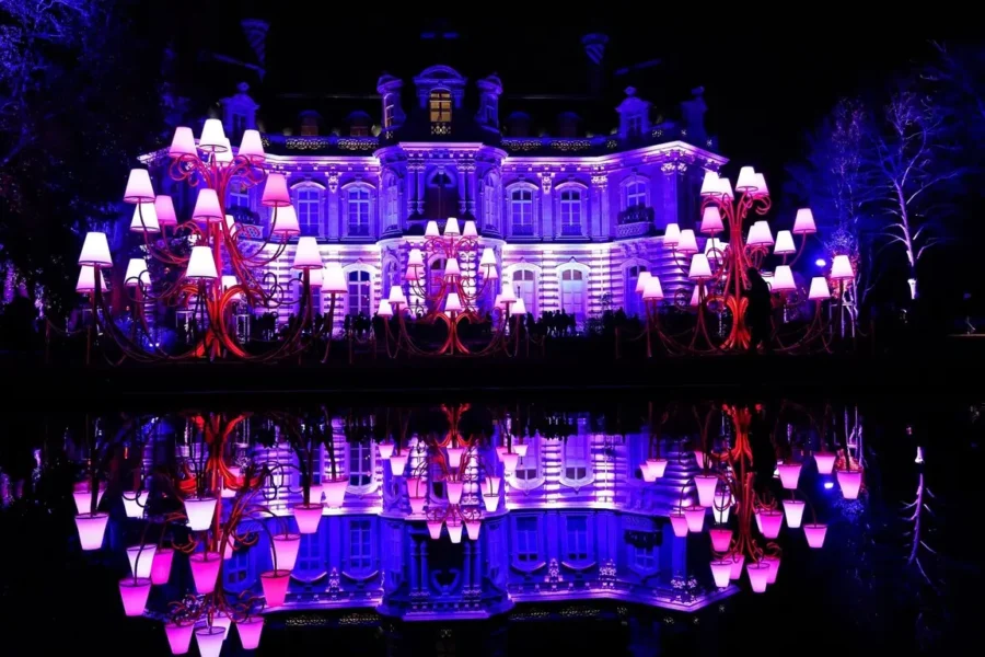 Violet lit historic mansion with oversized lamp sculptures and reflective pool at night.