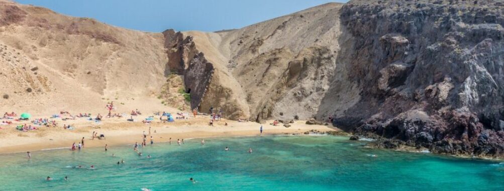 Turquoise volcanic cove beach with golden sand, cliffs, swimmers, umbrellas, and families relaxing.