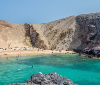 Turquoise volcanic cove beach with golden sand, cliffs, swimmers, umbrellas, and families relaxing.