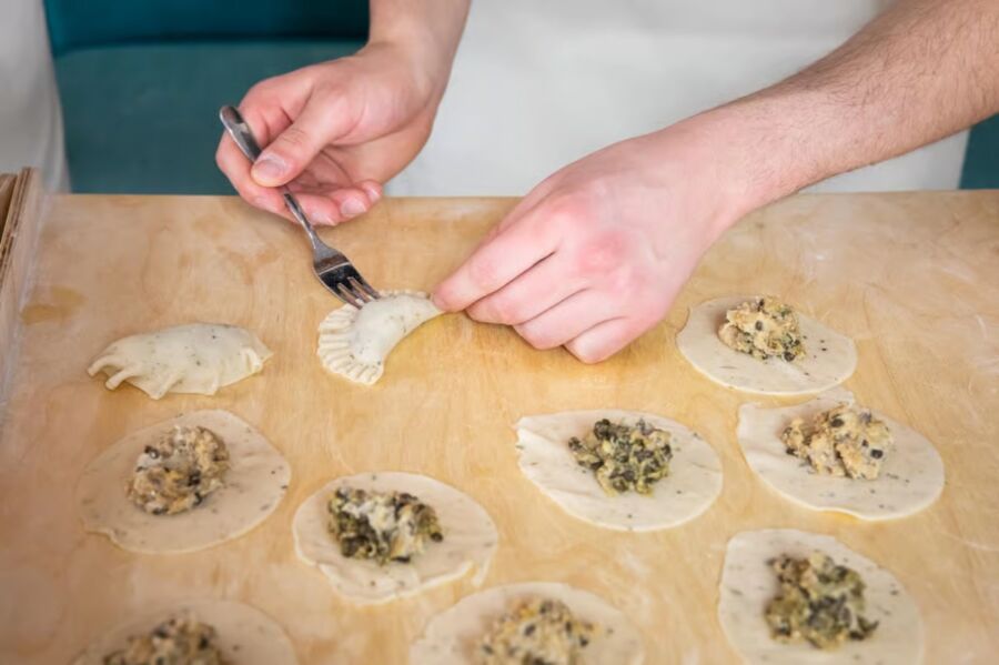 Person prepares dumplings on a wooden board in a Warsaw cooking class, enjoying drinks and the culinary experience.