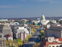 Aerial view of Washington, D.C., featuring the U.S. Capitol building centered in the background. The wide Pennsylvania Avenue is lined with autumn trees.