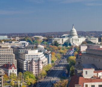Aerial view of Washington, D.C., featuring the U.S. Capitol building centered in the background. The wide Pennsylvania Avenue is lined with autumn trees.