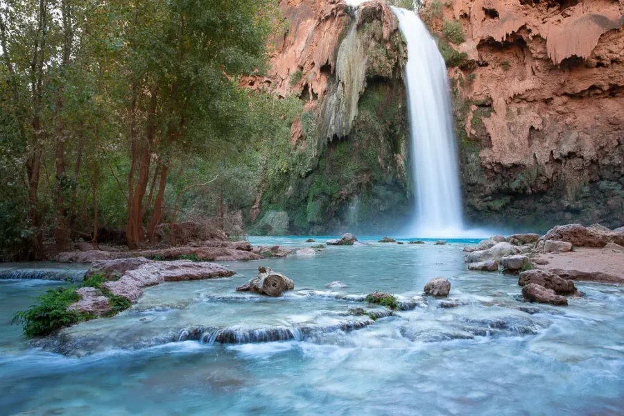 Majestic waterfall cascades into turquoise pool amid lush greenery and rugged cliffs.