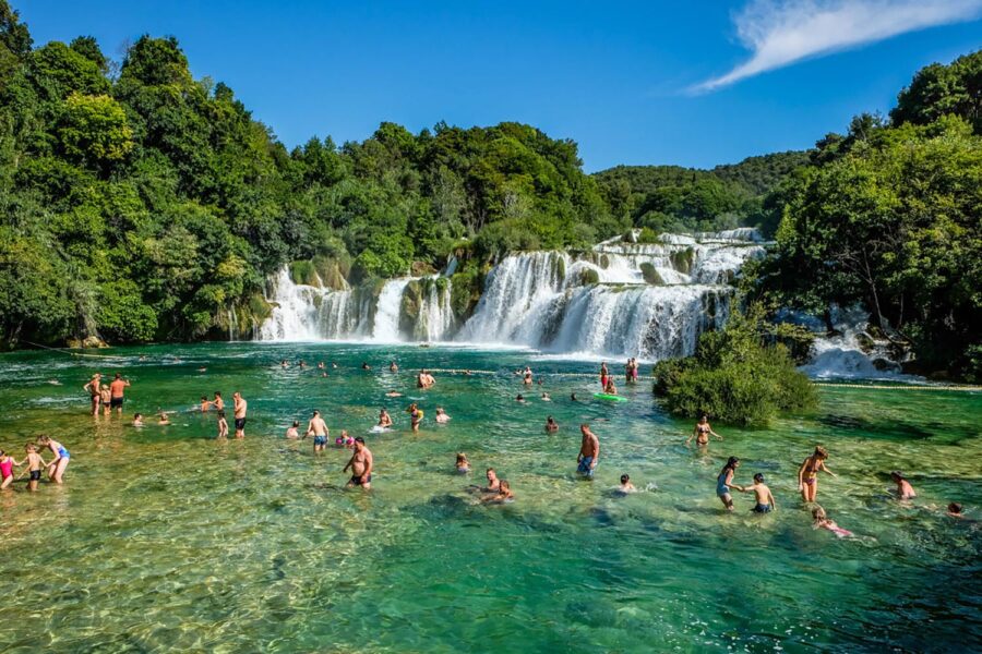 Scenic waterfall oasis with swimming, lush greenery, sunlight, happy visitors, and turquoise pool.