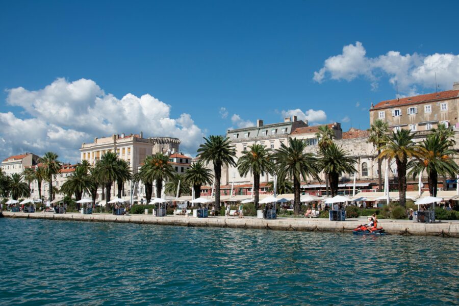 Coastal promenade with palm trees, cafes, azure waters, and dynamic jet ski action.