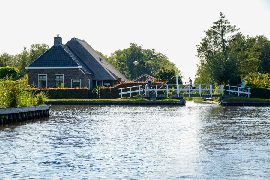 Scenic view of a Dutch house next to a canal, featuring a boat and cyclists in summer at National Park Weerribben-Wieden.