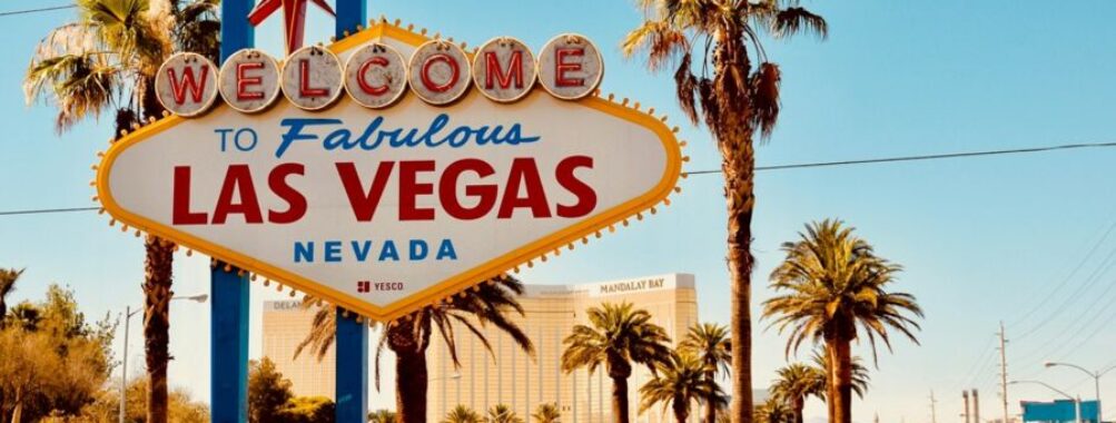Iconic Las Vegas sign with palm trees and Mandalay Bay, under a clear blue sky.