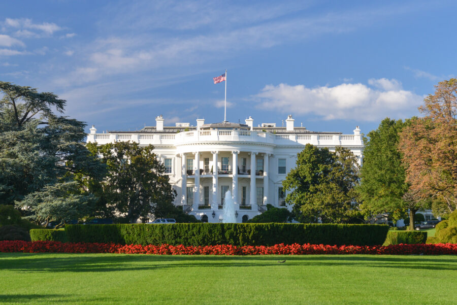 The White House, an iconic white mansion, stands majestically under a clear blue sky. Lush green lawns and vibrant red flowers enhance its grandeur.