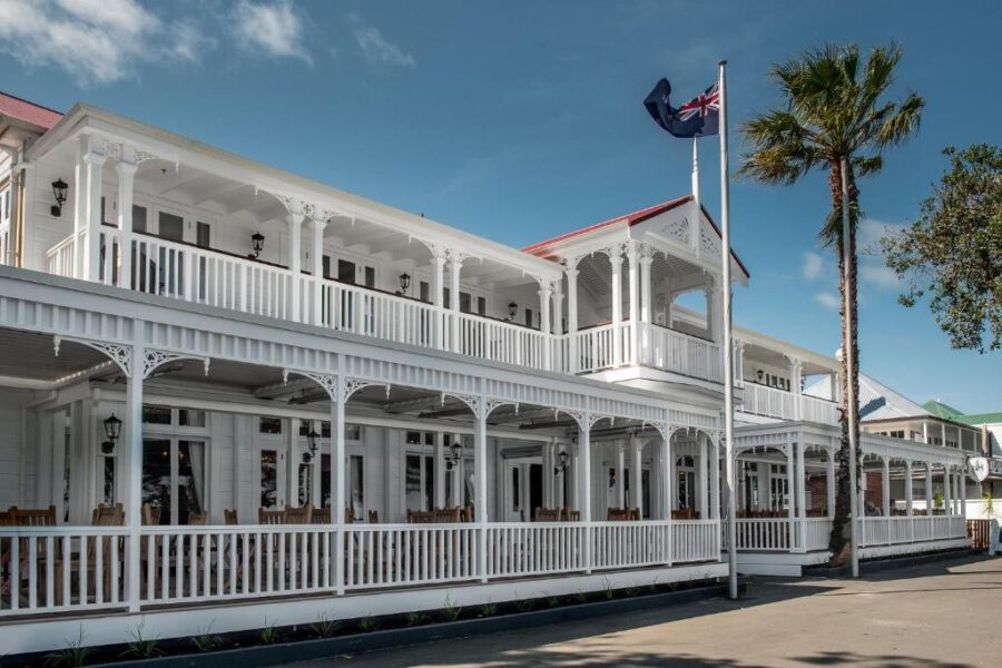 Historic white colonial seaside hotel with double verandas, palm tree, and New Zealand flag.