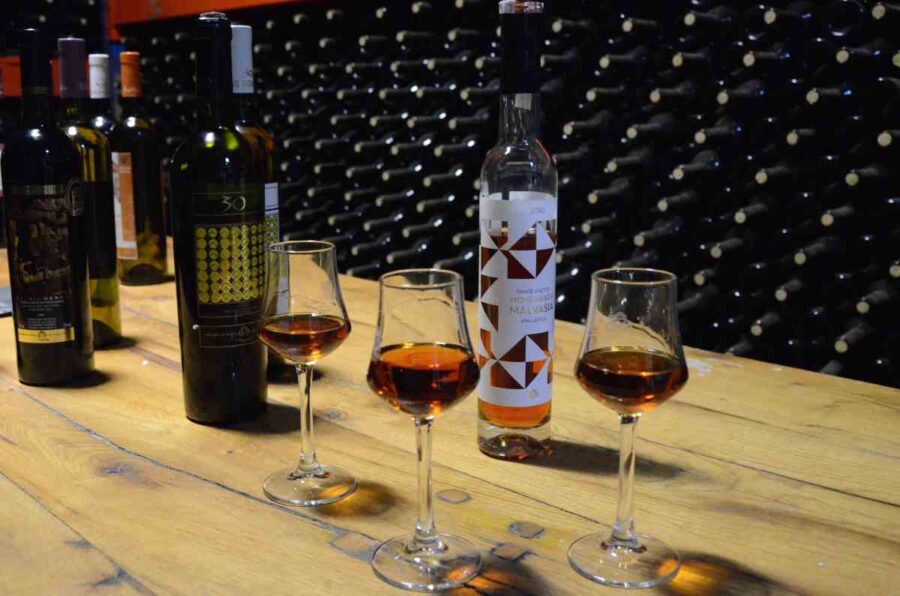 Malvasia wine tasting glasses with amber pours and bottles on rustic wooden table, cellar backdrop.