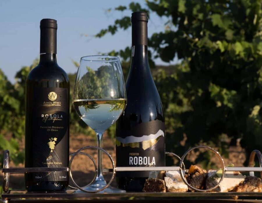 Robola wine bottles and glass on tray, vineyard background, featuring cheese and pastries.