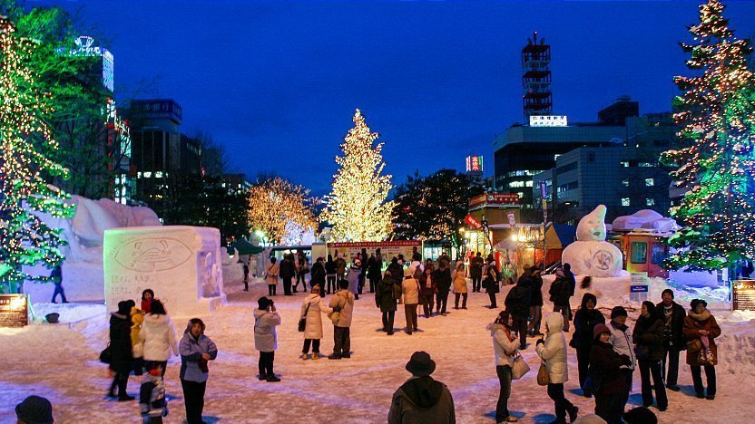 Snow festival in Sapporo city plaza at night with ice sculptures, Christmas lights, and crowds.
