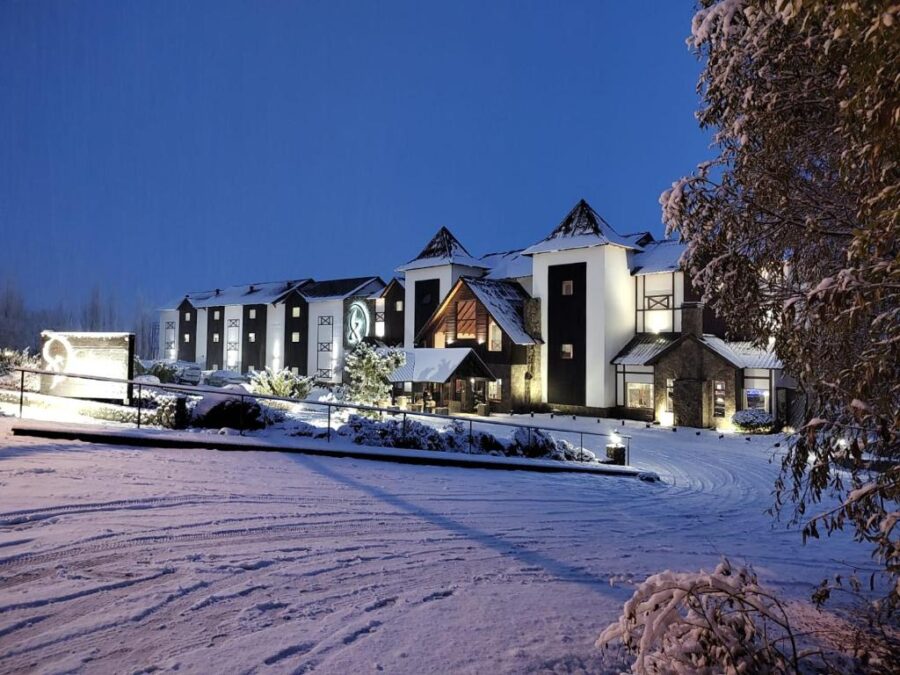 Snowy mountain hotel lodge at dusk with warm lights, winter resort driveway and forest backdrop.