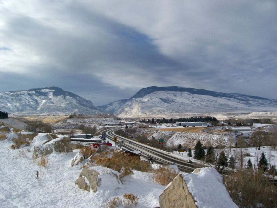 Snowy mountains and winding road with vehicles, evergreens, and buildings under a cloudy sky.