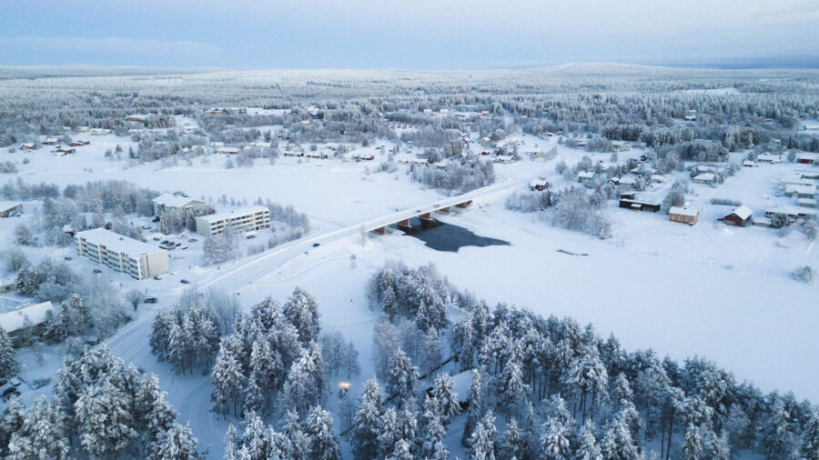 Aerial view of snowy winter landscape with river, bridge, and snow-covered evergreen trees.